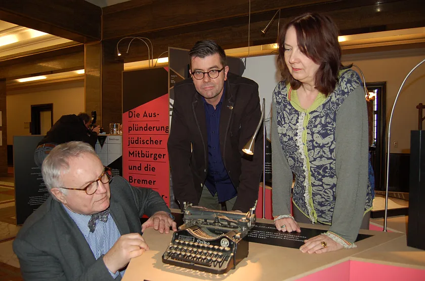 Finanzstaatsrat Henning Lühr, Dr. Jaromir Balcar und Bürgermeisterin Karolin Linnert (von links nach rechts) mit einer Schreibmaschine Typ „Erika“, die in der Ausstellung „Ausplündern und verwalten“ gezeigt wird. 20 solcher Schreibmaschinen wurden aus jüdischem Besitz beschlagnahmt und in der bremischen Finanzverwaltung bis mindestens 1952 genutzt.