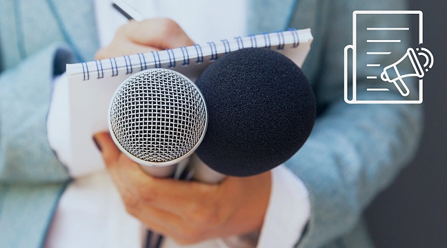 Symbolbild. Auf dem Bild sieht man vorne zwei Mikrofone und dahinter eine Person die etwas aufschreibt.