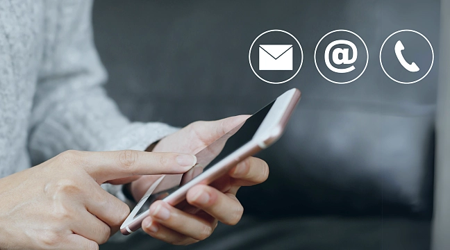 Symbolbild. Auf dem sieht man wie etwas in ein Smartphone getippt wird. Daneben werden Symbole Briefumschlag, ein @-Zeichen und ein Telefonhörer abgebildet.